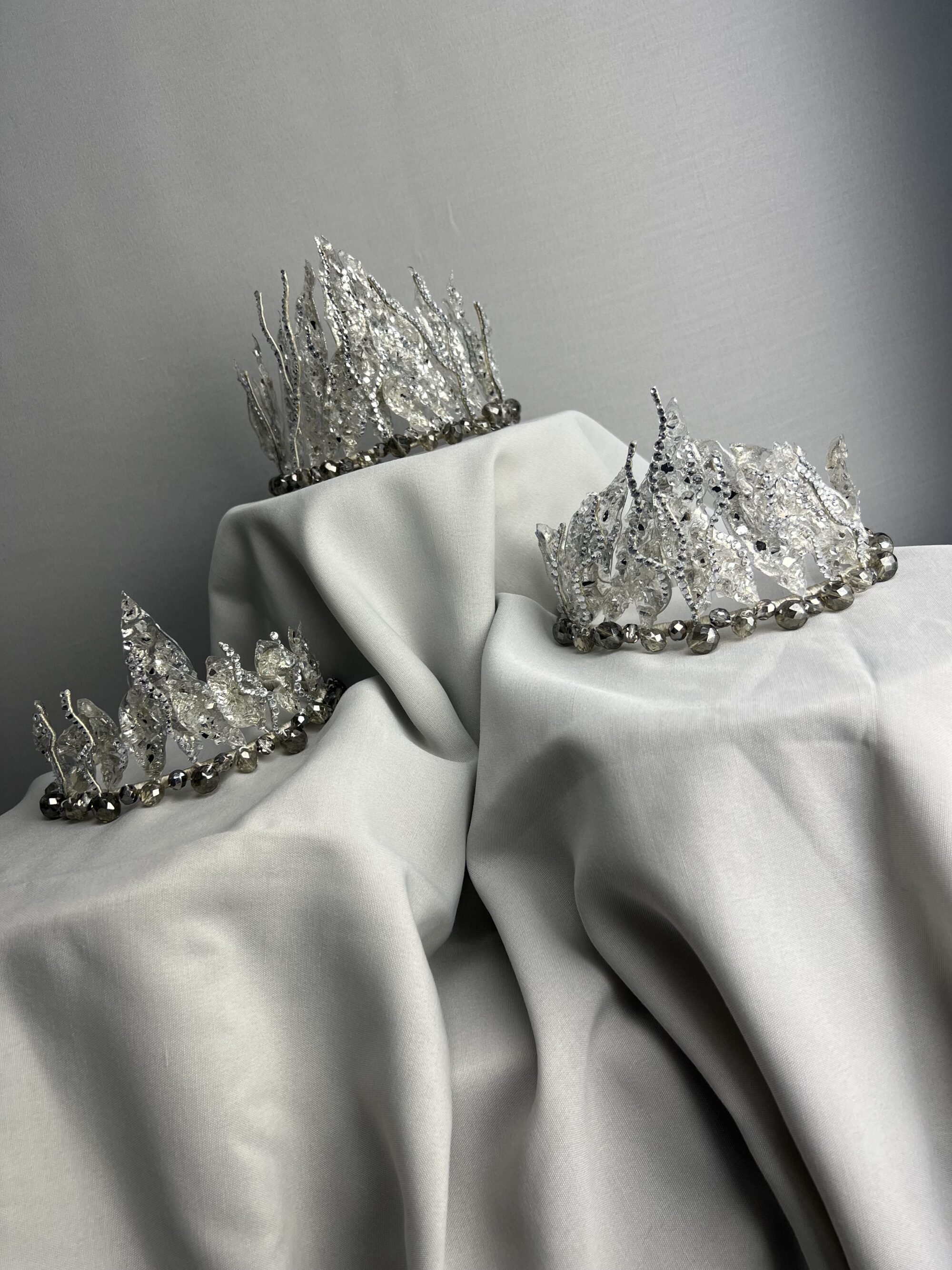 Tri strieborné a krištáľové svadobné šperky diadémy sú elegantne vystavené na nariasenej bielej látke na hladkom pozadí.