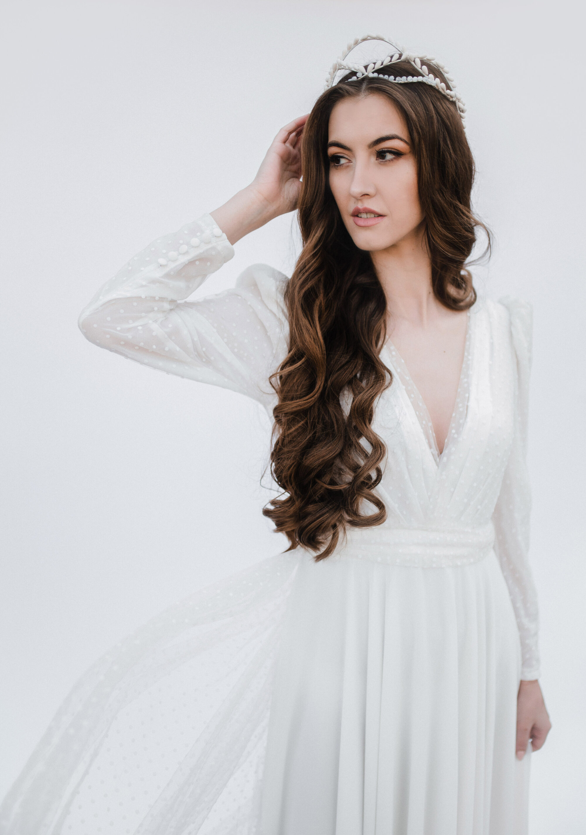 Žena v dlhých bielych šatách a čelenke, elegantná svadobná čelenka, stojí s rukou dotýkajúcou sa vlasov a pozerá na stranu na svetlom pozadí.
