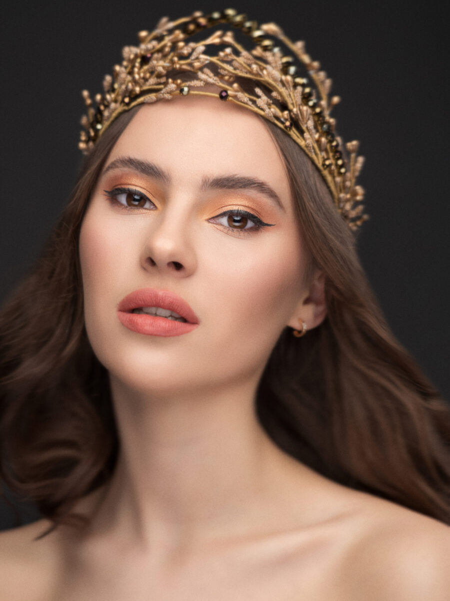 Žena s hnedými vlasmi má zlatú korunku, elegantný svadobný doplnok a jemný make-up na tmavom pozadí.