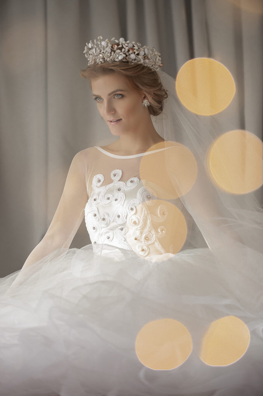 Nevesta v bielej róbe s kvetinovými detailmi, elegantne zdobená svadobnou korunkou a závojom, sedí na pozadí sivého závesu s jemnými bokeh svetlami v popredí.